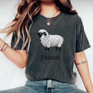 May include: Dark grey t-shirt featuring a detailed illustration of a sheep with black legs and face. The word "Found" is printed below the sheep in a simple, sans-serif font. The shirt has a vintage, worn look.