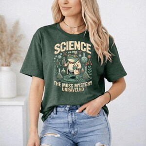 May include: Dark green t-shirt featuring the text "SCIENCE is my" and an illustration of a frog in a lab coat holding a test tube. The text "THE MOSS MYSTERY UNRAVELED" is below the illustration.