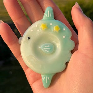 May include: A light mint green glass pendant shaped like a stylized sunfish. The piece features a small black eye, two yellow star accents, white dot details, and a central circular indentation with white line markings on the body surface.