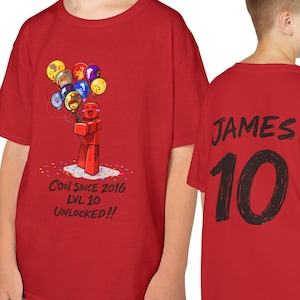 May include: Red t-shirt featuring a cartoon figure holding balloons. The front displays the text "Cool Since 2016 LVL 10 UNLOCKED!!". The back of the shirt has the name "JAMES" and the number "10" in black.