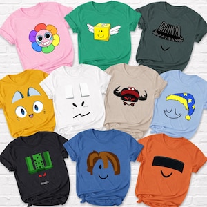 Puede incluir: Una colección de camisetas gráficas coloridas. Los diseños incluyen una flor sonriente, un cubo alado, una cara con sombrero, una cara de gato, una cara con rectángulos, un toro, una cara durmiente con gorro de dormir, un diseño de dominó, una cara con pelo castaño y una cara con una banda negra.