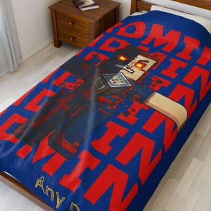 Puede incluir: Una colcha azul con un diseño gráfico rojo y negro. El diseño presenta un personaje pixelado sosteniendo un dispositivo, con las palabras "TOMMYINNIT" en rojo. También son visibles las palabras "Any D".