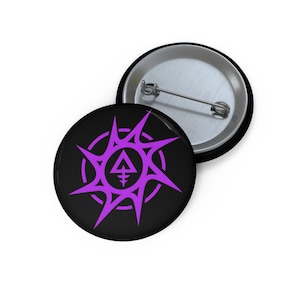 May include: Two black circular badges with a purple design. The design features a central triangle with a cross-like symbol, surrounded by a circle and spiky starburst shapes. One badge is face up, and the other is face down.