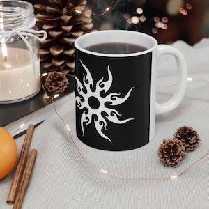 Puede incluir: Taza de café de cerámica negra con un diseño de sol blanco. La taza tiene un asa blanca y contiene un líquido oscuro. La taza está sobre una superficie gris claro con elementos decorativos.