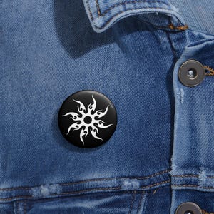 May include: A black button badge featuring a white tribal sun design. The round badge is set against a blue denim jacket.