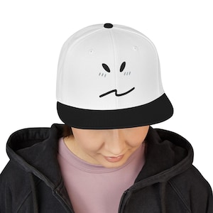 Puede incluir: Gorra de béisbol blanca con visera negra. La gorra presenta un diseño de rostro minimalista con ojos negros y una boca curva. La gorra es de estilo plano.