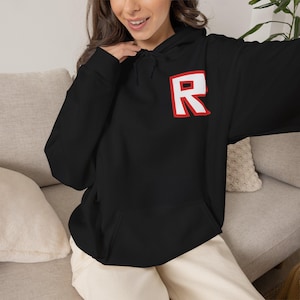 May include: A black long-sleeve hooded sweatshirt featuring a large red and white letter R graphic on the upper left chest. The hoodie includes a drawstring hood and a front kangaroo pocket, constructed from a solid black fabric material.