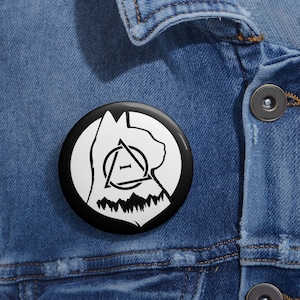 May include: A black and white button pin featuring a cat silhouette, a geometric design, and a mountain range. The pin is round and has a glossy finish. The pin is attached to a denim jacket.