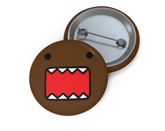 Puede incluir: Botón marrón con un personaje de dibujos animados con una boca roja abierta, dientes blancos y ojos negros. El botón es redondo y tiene un respaldo de alfiler metálico.