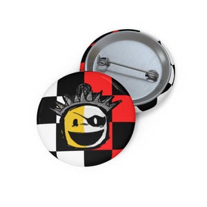 May include: Two round buttons with a graphic design. One button features a smiling face with a crown and eye patch on a red, black, and white checkered background. The other button has a black and white checkered pattern.