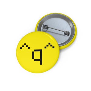 Puede incluir: Dos botones redondos amarillos con un diseño de cara pixelada. La cara tiene cejas pixeladas negras y una boca cuadrada negra. Un botón está boca arriba y el otro boca abajo, mostrando el alfiler.