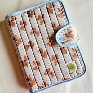 Pode incluir: Uma capa de almofada de mudança acolchoada com um padrão repetido de ursinhos de pelúcia castanhos sobre um fundo listrado creme e branco. A almofada tem uma borda azul clara e um fecho de pressão a condizer. Uma pequena etiqueta diz "RIRI MADE".
