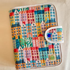 Può includere: Custodia in tessuto trapuntato con un colorato motivo a paesaggio urbano. Il design include file di case in vari colori come rosso, blu e giallo, con dettagli di finestre bianche. La custodia ha un bordo bianco e una chiusura a scatto con un bottone blu.