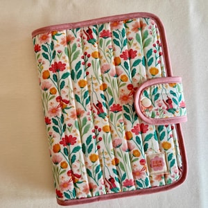 Può includere: Copertura trapuntata con stampa floreale, rifinitura rosa e chiusura con bottone abbinata. La copertura presenta uno sfondo bianco con un motivo di fiori rossi, rosa e gialli e foglie verdi. L'etichetta recita "RIBI MADE".