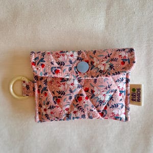 Könnte beinhalten: Kleine, gesteppte Tasche mit floralem Muster in Rosa, Weiß und Rot. Sie hat einen hellblauen Knopfverschluss und einen cremefarbenen Ring. Das Etikett lautet "lili MADE".