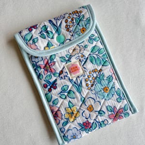 Könnte beinhalten: Eine gesteppte Hülle mit floralem Muster, hellblauer Einfassung und Knopfverschluss. Die Hülle hat einen weißen Hintergrund mit einem Muster aus bunten Blumen und grünen Blättern. Das Etikett lautet "RIRI MADE".