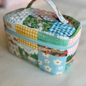 May include: A small, rectangular quilted cosmetic bag with a handle. The bag features a patchwork design with various patterns, including gingham, floral, and scenes with rabbits. It has a light blue zipper and a matching handle.