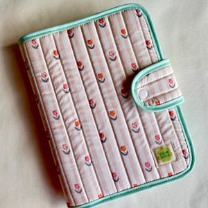 May include: A quilted, pink and white striped cover with a floral tulip pattern. The cover has a mint green border and a matching closure with a white button. A small label reads "Riri Made".