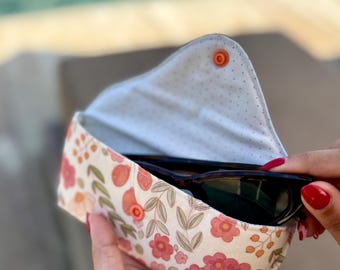 May include: Floral print sunglasses case with a snap closure. The case features a cream background with pink and orange flowers and green leaves. The interior is light blue with small white polka dots. The label reads "RIRI MADE".