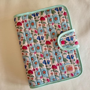 May include: A quilted floral notebook cover with a light blue border and a matching closure. The cover features a pattern of blue and red flowers on a cream background. The closure has a pink button. The label reads "SIRIRI MADE".