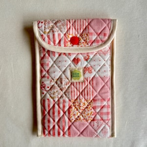 May include: A quilted, pink and white fabric case with a flap closure. The case features a patchwork design with various patterns, including stripes and floral prints. A small red button secures the flap. The label reads "GIGI MADE".