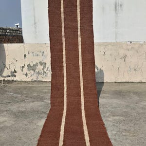 May include: A long, rectangular runner rug featuring a deep brown, textured woven surface. The runner is decorated with two parallel, vertical off-white stripes that run the entire length of the piece. The edges show a slightly frayed, natural finish.