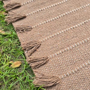 May include: A brown woven rug with white stripes and decorative tassels along the edge. The rug is made of a textured material and is placed on green grass. The tassels are a darker shade of brown.