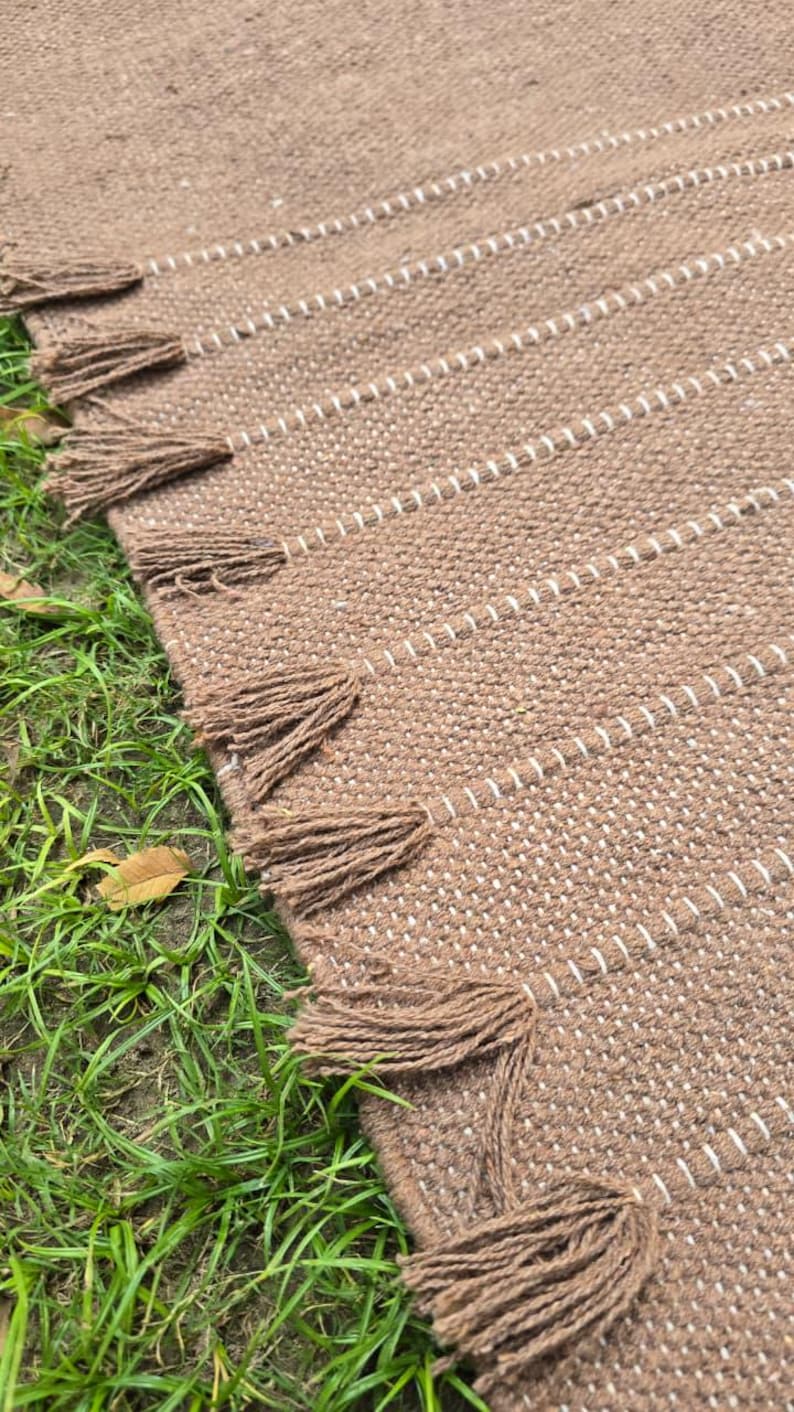 May include: A brown woven rug with white stripes and decorative tassels along the edge. The rug is made of a textured material and is placed on green grass. The tassels are a darker shade of brown.