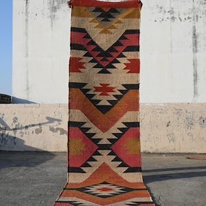 以下が含まれることがあります： 幾何学模様の長方形の丸められたラグ。赤、黒、タン、金の三角形とひし形の繰り返し模様が特徴です。白い壁を背景に屋外に展示されています。