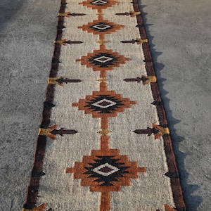 May include: A long, rectangular rug with a geometric pattern in shades of brown, black, and beige. The rug features repeating diamond and arrow motifs, with a dark brown border and fringe at the ends. The rug is made of a woven material.