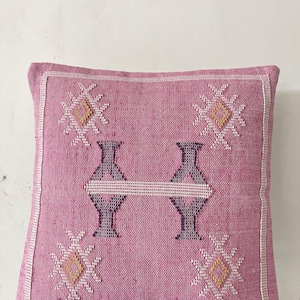 May include: A square, pink decorative pillow with a geometric design. The pillow features a central pattern of two stylized vases connected by a horizontal line, and four starburst-like designs. The edges are trimmed with white.