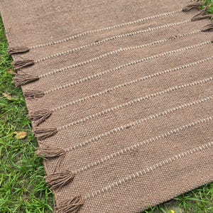 May include: A rectangular brown woven rug with white stitching and brown tassels. The rug is lying on green grass. The rug has a textured appearance and is likely made of natural fibers.