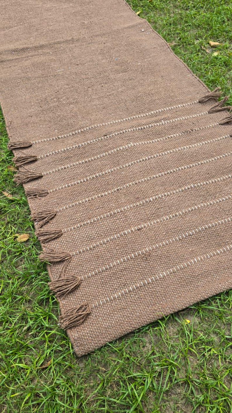 May include: A rectangular brown woven rug with white stitching and brown tassels. The rug is lying on green grass. The rug has a textured appearance and is likely made of natural fibers.