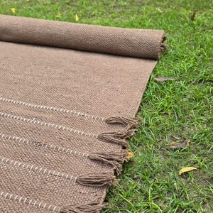 May include: A rolled-up brown woven rug with white stripes and tassels, resting on green grass. The rug is made of a textured material and has a rectangular shape. The tassels are at the end of the rug.