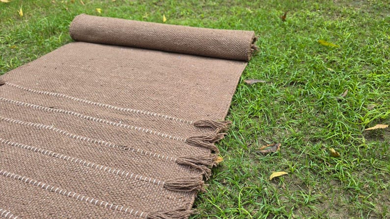 May include: A rolled-up brown woven rug with white stripes and tassels, resting on green grass. The rug is made of a textured material and has a rectangular shape. The tassels are at the end of the rug.
