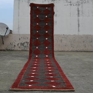 May include: A long, rectangular runner rug with a geometric diamond pattern. The rug features a dark red border and diamond shapes, with gray diamond shapes in the center. White rectangular accents are between the red diamonds.