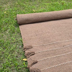 May include: A rolled-up brown woven rug with white stripes and tassels. The rug is made of a textured material and is partially unrolled on a grassy surface. The tassels are a darker shade of brown.