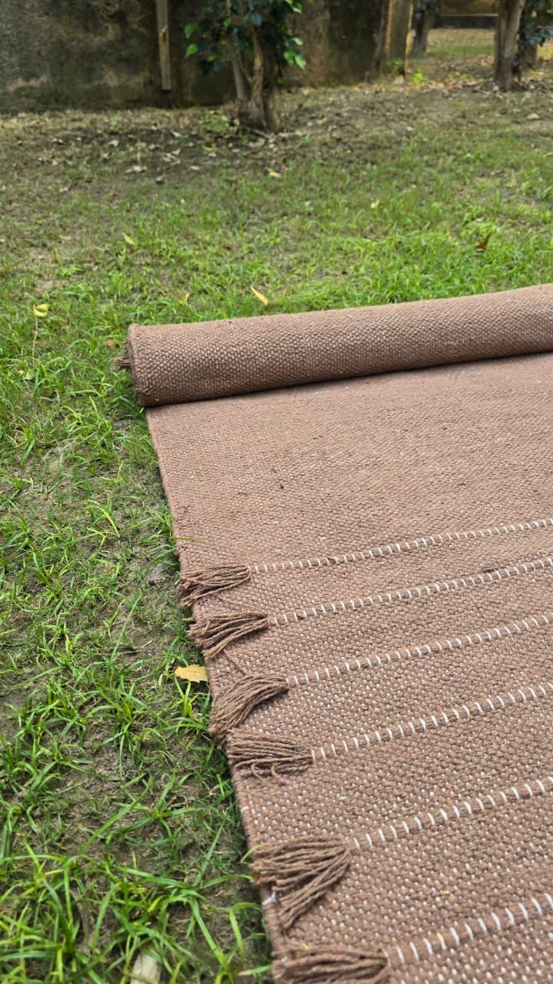 May include: A rolled-up brown woven rug with white stripes and tassels. The rug is made of a textured material and is partially unrolled on a grassy surface. The tassels are a darker shade of brown.