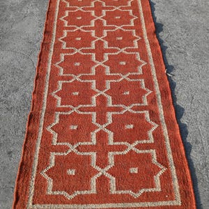 Puede incluir: Una alfombra rectangular de color óxido con un patrón geométrico en crema. El diseño presenta formas de estrella y cruz repetidas. La alfombra tiene flecos en ambos extremos. Adecuada para un pasillo o entrada, mide aproximadamente 200 cm de largo.