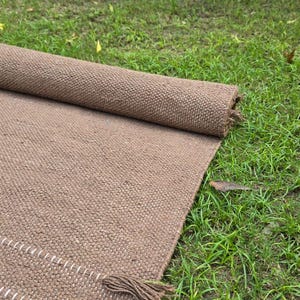 May include: A rolled-up brown woven rug lies partially unrolled on green grass. The rug has a textured surface and a fringe detail. The unrolled portion shows a white stitched border. The rug is made of a natural fiber.