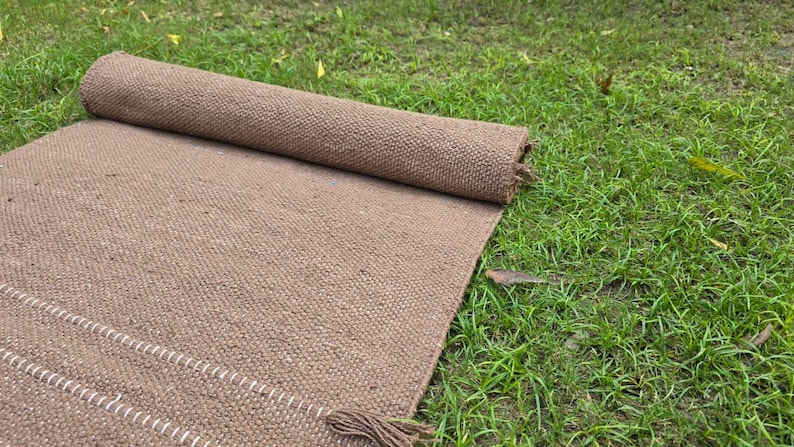 May include: A rolled-up brown woven rug lies partially unrolled on green grass. The rug has a textured surface and a fringe detail. The unrolled portion shows a white stitched border. The rug is made of a natural fiber.