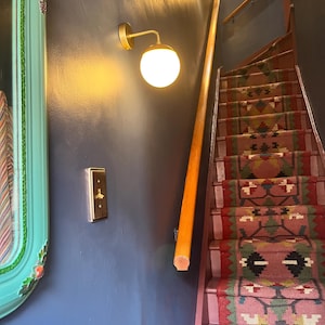 Pu&ograve; includere: Una scala con un tappeto a motivi geometrici rosa, verdi e rossi. Una parete blu scuro presenta un interruttore a levetta color ottone e una lampada da parete con paralume sferico in vetro bianco e braccio color ottone.