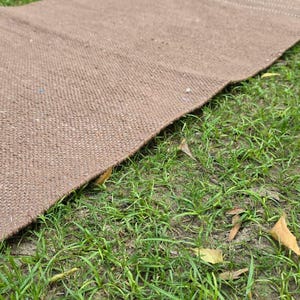 May include: A brown woven rug lies on a bed of green grass. The rug has a textured surface and a rectangular shape. The rug is made of a natural fiber and has a simple, rustic design.