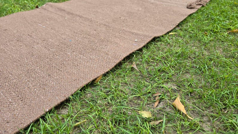 May include: A brown woven rug lies on a bed of green grass. The rug has a textured surface and a rectangular shape. The rug is made of a natural fiber and has a simple, rustic design.