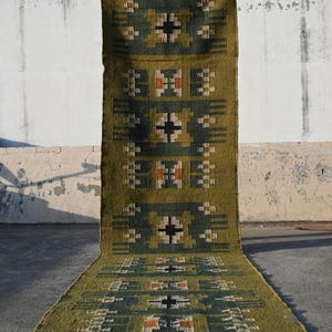 Pode incluir: Um tapete longo verde-oliva com um padrão geométrico em verde escuro, preto e laranja. O tapete é exibido verticalmente, mostrando seu comprimento e design. O padrão inclui quadrados e cruzes.