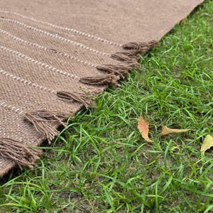 May include: A brown woven rug with white stripes and tassels along the edge. The rug is partially laid out on green grass. The rug's texture is visible, and the tassels are a darker brown.
