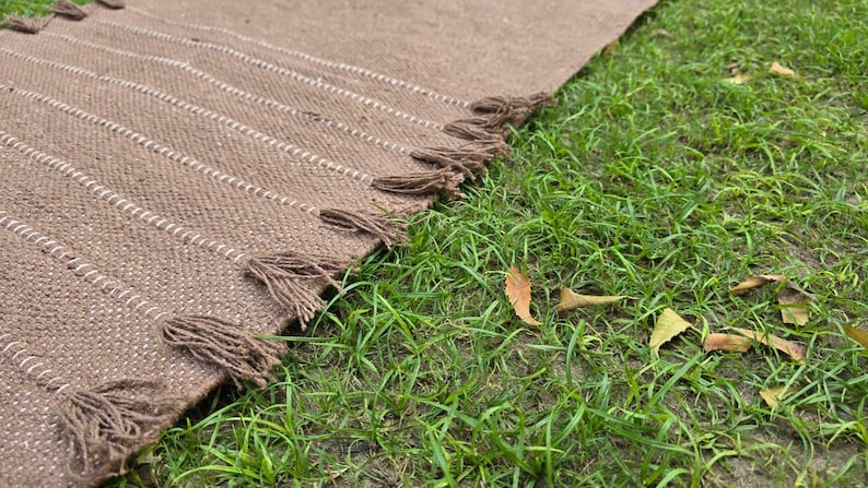 May include: A brown woven rug with white stripes and tassels along the edge. The rug is partially laid out on green grass. The rug's texture is visible, and the tassels are a darker brown.