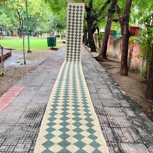 May include: A long, rectangular rug with a repeating diamond pattern in teal and beige. The rug has a fringed edge and is displayed outdoors on a tiled walkway, showcasing its length and design.