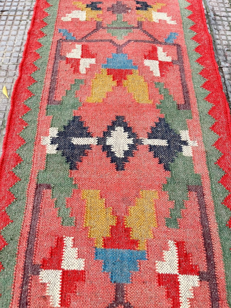 Pu&ograve; includere: Un tappeto passatoia rettangolare con base rosso corallo e un motivo geometrico nei toni del blu scuro, giallo senape, verde salvia e bianco. Il bordo presenta un profilo a zig-zag dentellato in rosso e verde che incornicia i motivi.