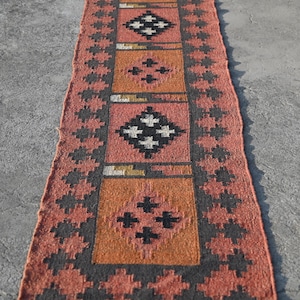 Könnte beinhalten: Ein langer, rechteckiger Läufer mit einem sich wiederholenden geometrischen Muster aus schwarz-weißen Rauten, die in abwechselnden orangefarbenen und terrakottafarbenen Quadraten zentriert sind, umrandet von einer dunkelgrauen Zackenkante und dunklen Fransen.